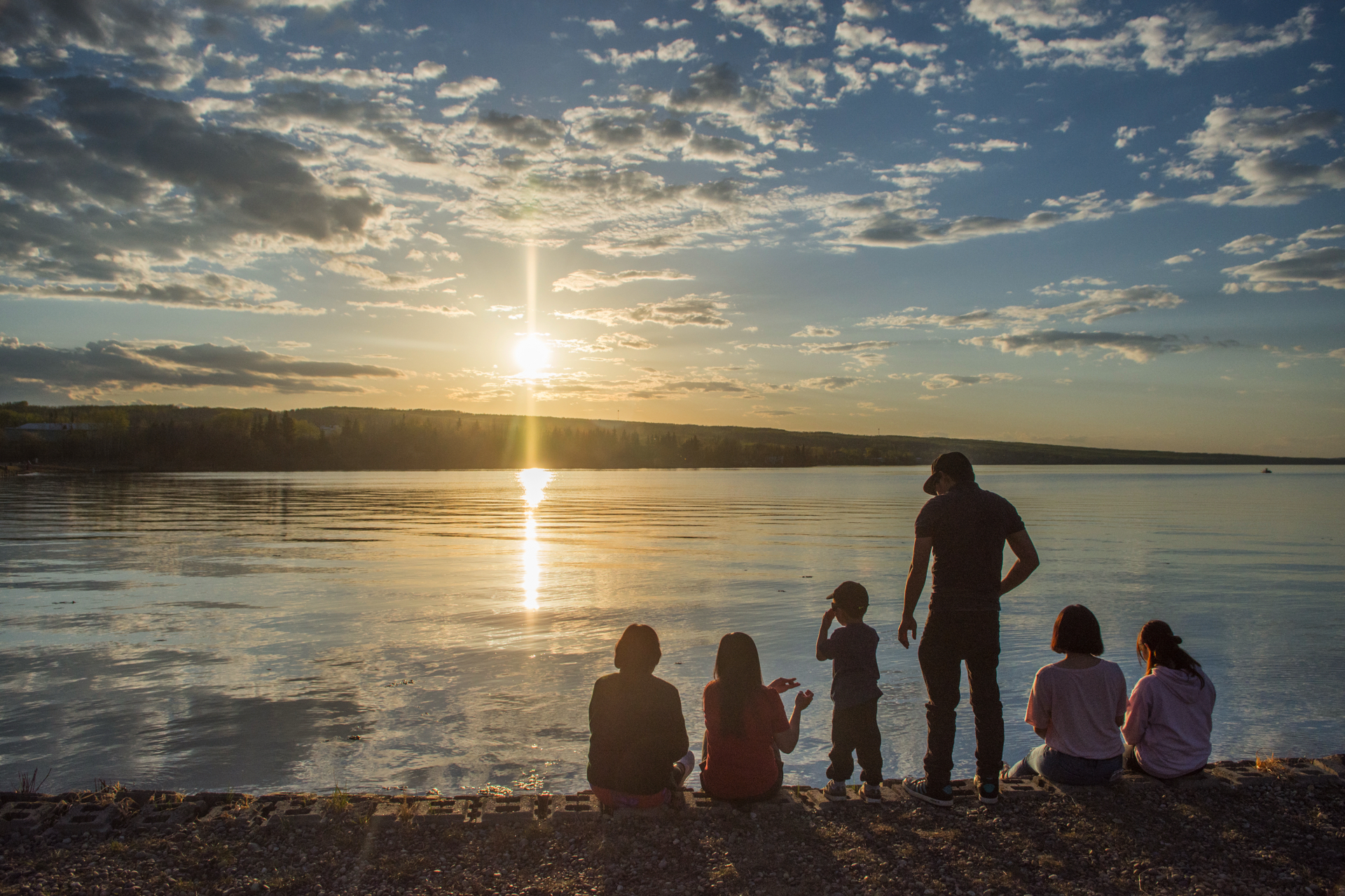 A group of people of varying ages are gathered on the edge of a lake, watching the sun set