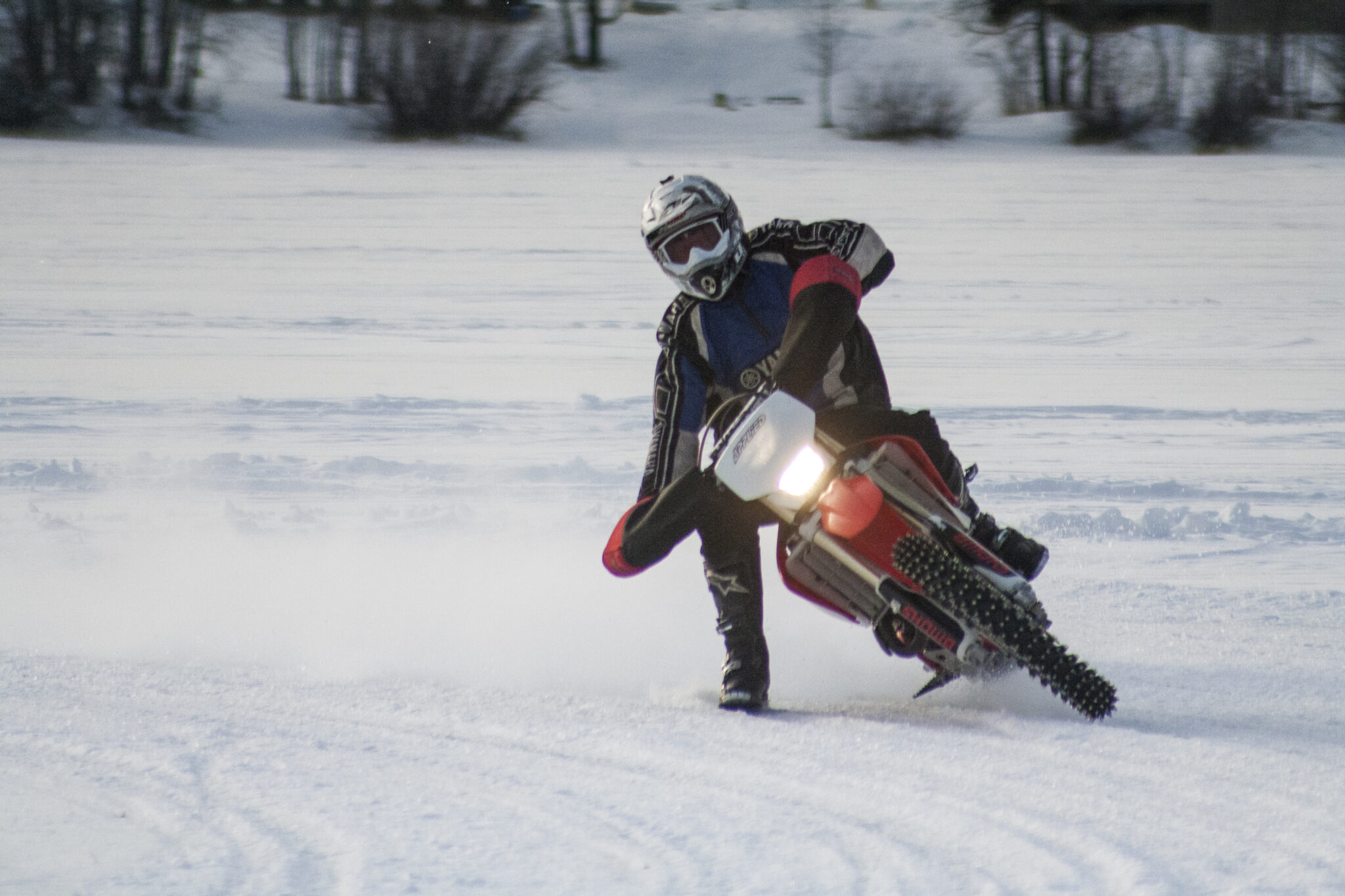 A person is riding a motocross motorcycle around a corner, sliding in the snow. The bike has been outfitted with special studded tires and "gloves" that attach directly to the handlebars.
