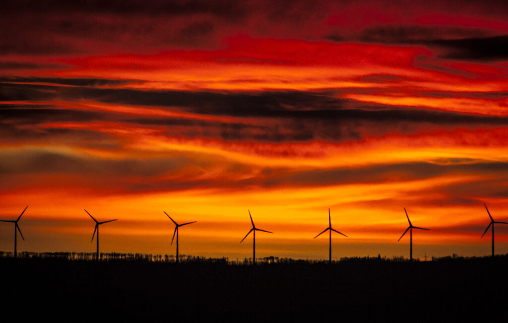 A row of wind turbine generators along a ridge are shown in silhouette against a bright red and orange sunset