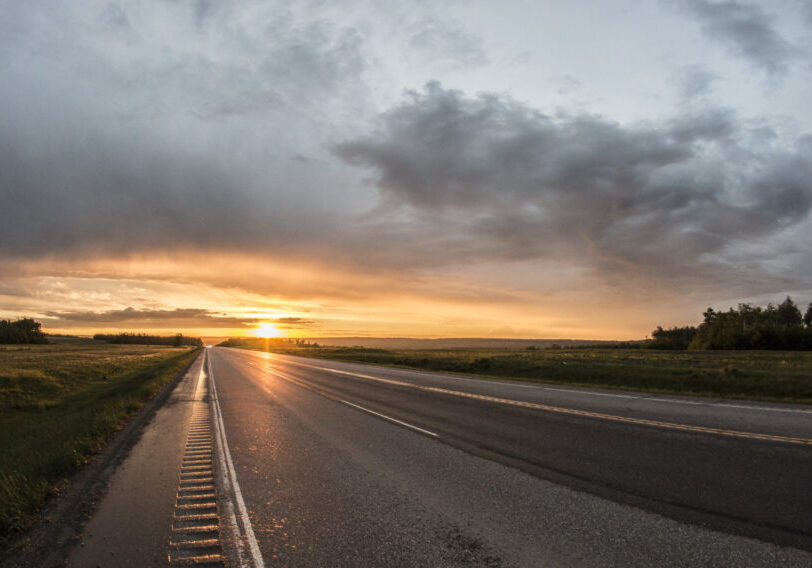 A highway in the South Peace, with no traffic. The sun is shining on the horizon casting a golden glow over the scene.