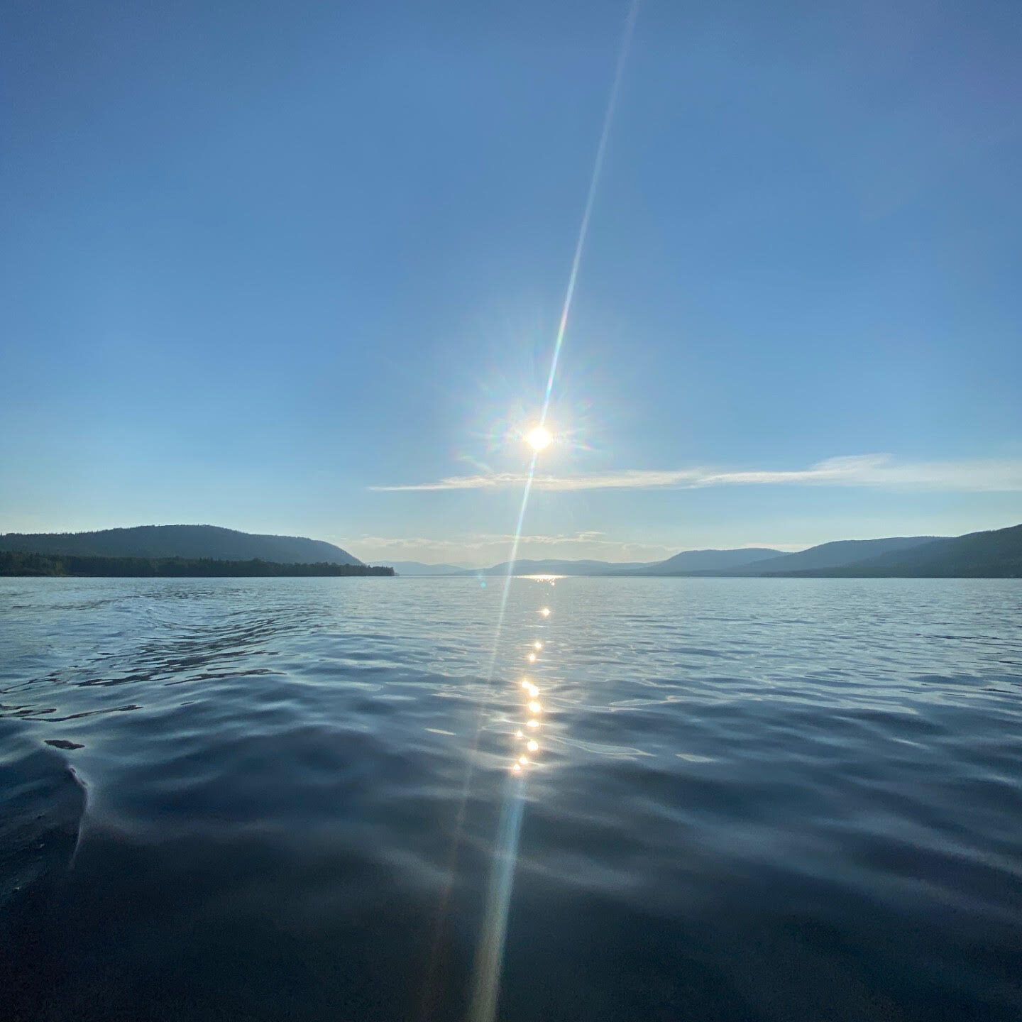 A view across the serene waters of Moberly Lake towards the sun, low in a vivid blue sky