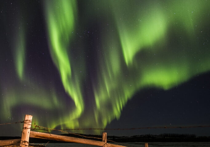 Northern lights lighting up the sky in bright streaks of green above a rustic rural barbed wire fence.