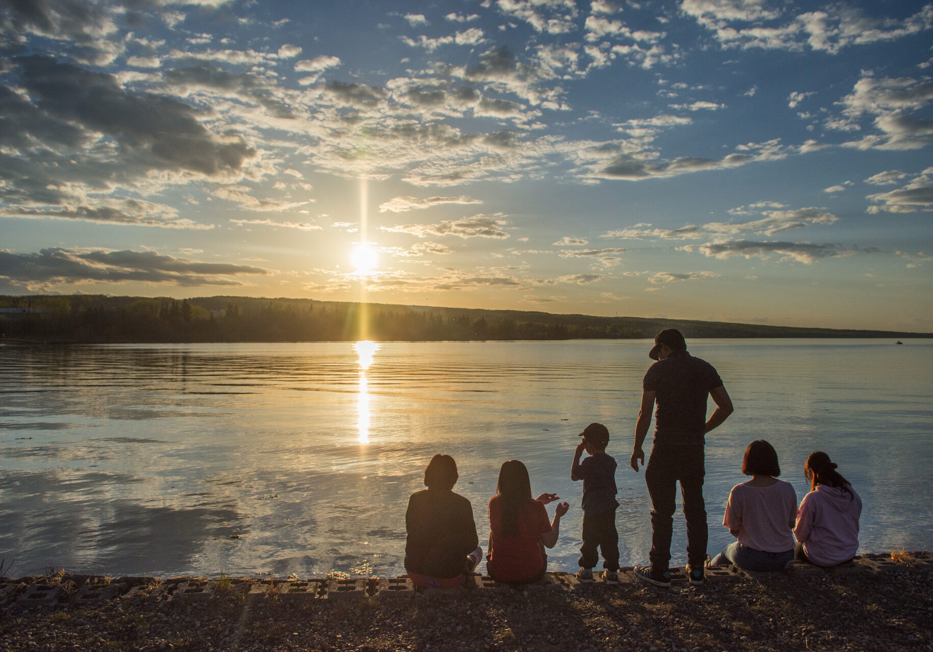 A group of people of varying ages are gathered on the edge of a lake, watching the sun set