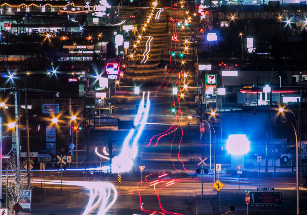 A shot of Dawson Creek at night, looking down 8th Street. Many bright lights from signage can be seen, along with long glowy red and white streaks from the vehicle traffic going up and down the street.